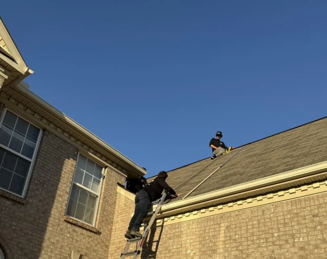 Three men are on a brick house. Two men are on the roof and one is standing by a ladder at the side of the house.